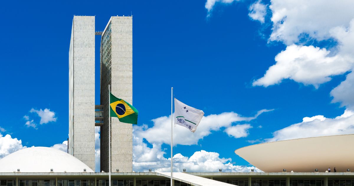 A imagem retrata o Congresso Nacional do Brasil, situado em Brasília. No centro, destacam-se duas torres retangulares e altas, que compõem os edifícios principais. À esquerda das torres, vê-se uma cúpula branca, representando a Câmara dos Deputados, enquanto à direita há uma estrutura em forma de prato invertido, que corresponde ao Senado Federal. Em primeiro plano, estão duas bandeiras hasteadas: a bandeira do Brasil à esquerda e a bandeira do Mercosul à direita. O céu ao fundo está azul com nuvens brancas esparsas, compondo um cenário claro e iluminado. (governo e ciber)