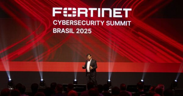 Uma pessoa está em pé sobre um palco, apresentando durante o evento "Fortinet Cybersecurity Summit Brasil 2025". Ao fundo, há uma tela grande com o nome e o ano do evento em texto branco, sobre um design abstrato em vermelho e preto. Na frente, vê-se o público sentado, voltado para o palco.