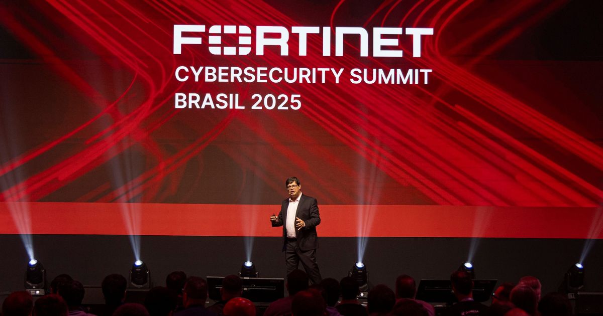 Uma pessoa está em pé sobre um palco, apresentando durante o evento "Fortinet Cybersecurity Summit Brasil 2025". Ao fundo, há uma tela grande com o nome e o ano do evento em texto branco, sobre um design abstrato em vermelho e preto. Na frente, vê-se o público sentado, voltado para o palco.