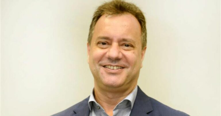 Silvio Ferraz de Campos, CEO da Positivo Servers & Solutions está em pé diante de um fundo claro, vestindo um terno azul escuro e uma camisa social cinza clara.