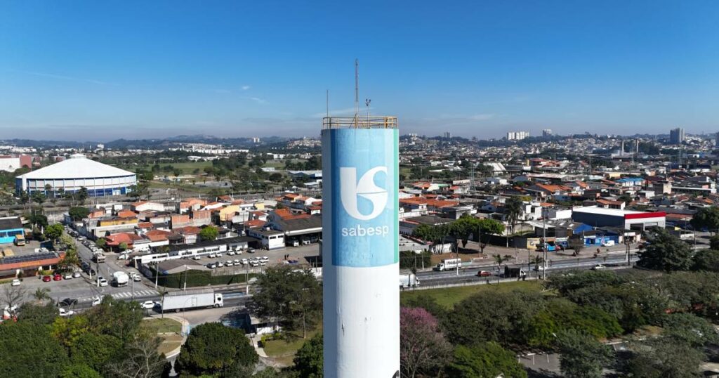 Vista aérea de uma região urbana com destaque para um reservatório cilíndrico branco da Sabesp (Companhia de Saneamento Básico do Estado de São Paulo), com o logotipo azul da empresa no centro. Ao fundo, há um bairro com casas, comércios e ruas movimentadas, além de um grande ginásio coberto à esquerda e edifícios espalhados ao longe. O céu está limpo e azul, indicando um dia ensolarado (inteligentes, Sabesprev)