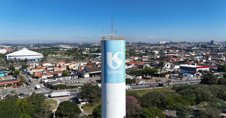 Vista aérea de uma região urbana com destaque para um reservatório cilíndrico branco da Sabesp (Companhia de Saneamento Básico do Estado de São Paulo), com o logotipo azul da empresa no centro. Ao fundo, há um bairro com casas, comércios e ruas movimentadas, além de um grande ginásio coberto à esquerda e edifícios espalhados ao longe. O céu está limpo e azul, indicando um dia ensolarado (inteligentes, Sabesprev)