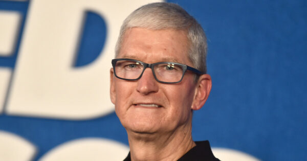 Tim Cook, CEO da Apple, aparece na imagem em um close, vestindo óculos de armação escura e camisa preta, com fundo azul e parte de uma inscrição branca ao fundo.