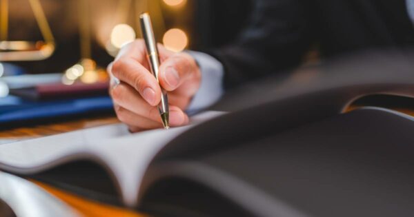 Pessoa de terno escrevendo com caneta dourada em um documento sobre a mesa, com balança de justiça desfocada ao fundo, representando gestão de contratos e assuntos jurídicos.