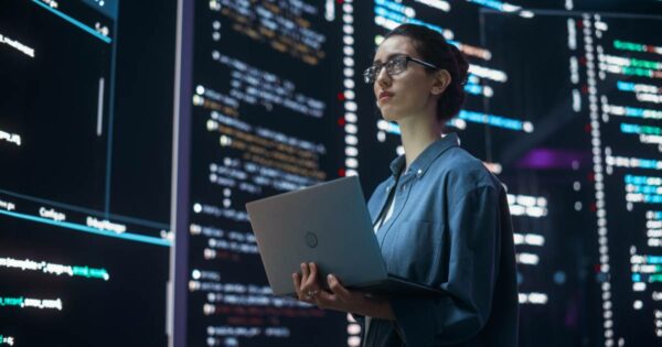 Mulher usando óculos e camisa azul, segurando um notebook, em frente a grandes telas com códigos de programação e dados iluminados (segurança, infraestrutura)