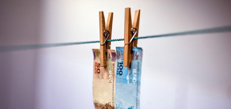 Imagem de duas cédulas de real brasileiro, uma de 50 e outra de 100 reais, presas por prendedores de madeira em um varal, sugerindo simbolicamente o conceito de lavagem de dinheiro.