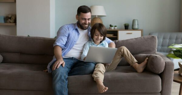 Pai e filho sentados juntos no sofá, sorrindo enquanto usam um notebook, em um ambiente de sala de estar iluminado e aconchegante (pais)
