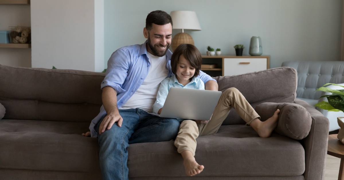 Pai e filho sentados juntos no sofá, sorrindo enquanto usam um notebook, em um ambiente de sala de estar iluminado e aconchegante (pais)