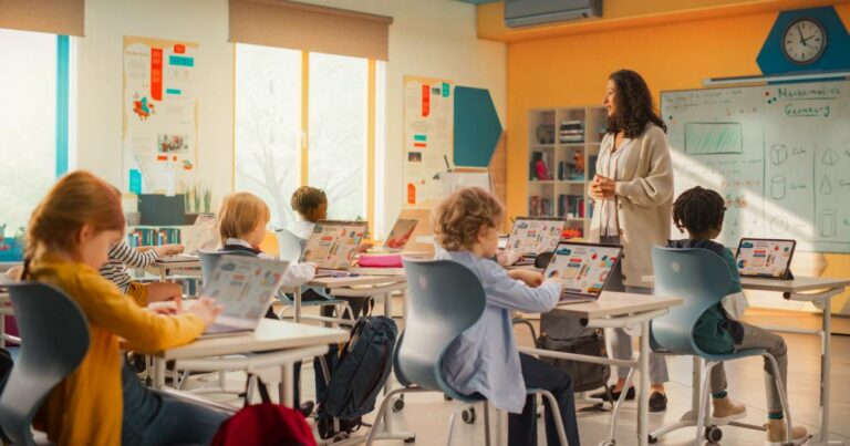 Sala de aula moderna com crianças usando laptops coloridos em suas carteiras, enquanto uma professora em pé, sorridente, conduz a atividade. Ao fundo, há um quadro branco com anotações de matemática e geometria, representando o uso de tecnologia na educação (Programa, cátedra)