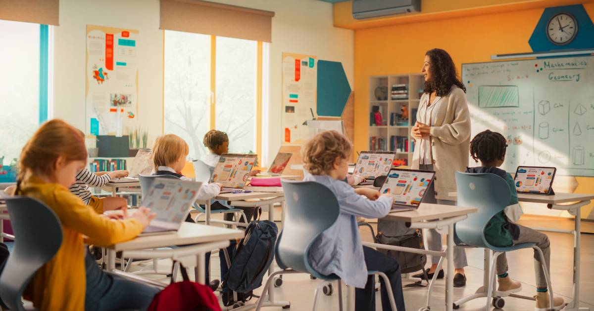 Sala de aula moderna com crianças usando laptops coloridos em suas carteiras, enquanto uma professora em pé, sorridente, conduz a atividade. Ao fundo, há um quadro branco com anotações de matemática e geometria, representando o uso de tecnologia na educação (Programa, cátedra)