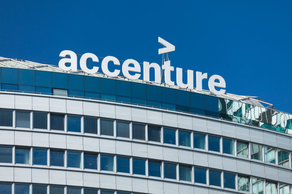 Parte superior de um edifício moderno com o logotipo da "accenture" exibido de forma proeminente no topo. O logotipo é composto pela palavra "accenture" em letras minúsculas, acompanhada de um símbolo de maior (>) acima dela. O prédio possui vários andares com grandes janelas, e o céu azul aparece ao fundo.