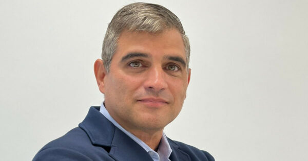 Homem de meia-idade, de cabelos curtos grisalhos, vestindo camisa azul clara e blazer azul escuro, em foto de retrato com fundo branco. Trata-se de Adriano Caldevilla, novo diretor de Tecnologia da Angola Cables.