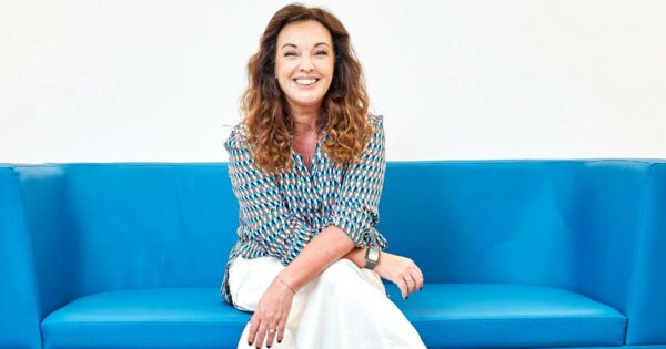 Carmela Borst, da Soul Code, sorridente sentada em um sofá azul, usando blusa estampada colorida e calça branca