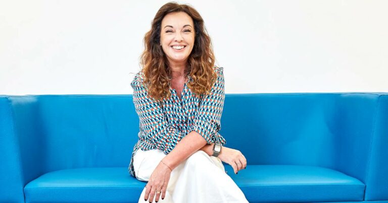 Carmela Borst, da Soul Code, sorridente sentada em um sofá azul, usando blusa estampada colorida e calça branca