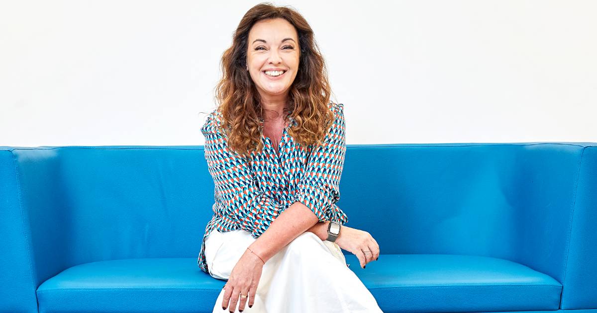 Carmela Borst, da Soul Code, sorridente sentada em um sofá azul, usando blusa estampada colorida e calça branca