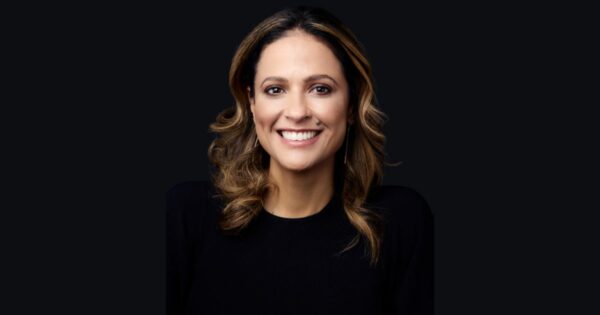 Mulher sorridente em foto de estúdio, com cabelo castanho ondulado e mechas mais claras, vestindo blusa preta. Fundo liso escuro. Trata-se de Alaine Charchat, CIO da Reckitt para a América Latina (Líder)