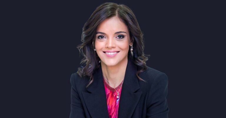 Mulher sorridente de cabelos castanhos e ondulados, usando blazer preto sobre blusa vermelha estampada, posando em frente a fundo escuro. Trata-se de Ana Paula Assis, vice-presidente sênior e presidente da IBM para Europa, Oriente Médio, África e Mercados em Crescimento.