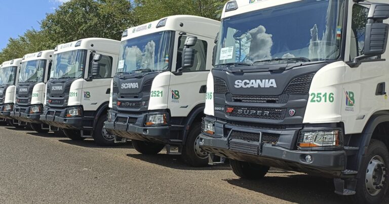 Fila de caminhões brancos da marca Scania, estacionados lado a lado, com a logo da empresa **I.B Logística e Transportes** estampada nas portas (reduz)