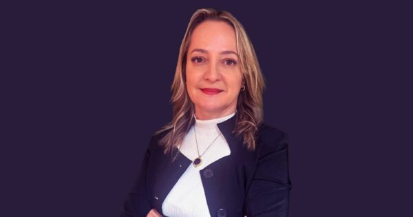 Foto de Angelica Vitali, head Brasil da T-Systems, vestindo blazer preto sobre blusa branca, colar com pingente redondo e posando com os braços cruzados sobre fundo roxo uniforme.