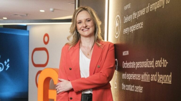 Mulher sorridente, de cabelos loiros e ondulados, veste blazer coral e blusa branca, posando em pé ao lado de uma parede com textos em inglês sobre propósito e missão da empresa. No fundo, há elementos de design corporativo com formas geométricas e iluminação moderna. A imagem mostra Amanda Andreone, vice-presidente de Vendas da Genesys no Brasil.