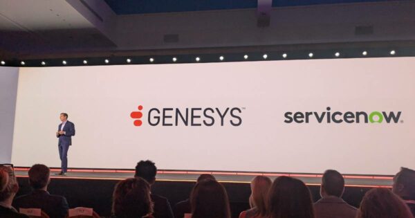 Homem de terno azul, identificado como Tony Bates, CEO da Genesys, está em um palco apresentando diante de um grande telão com os logotipos da Genesys e da ServiceNow. Na frente, várias pessoas assistem à apresentação.
