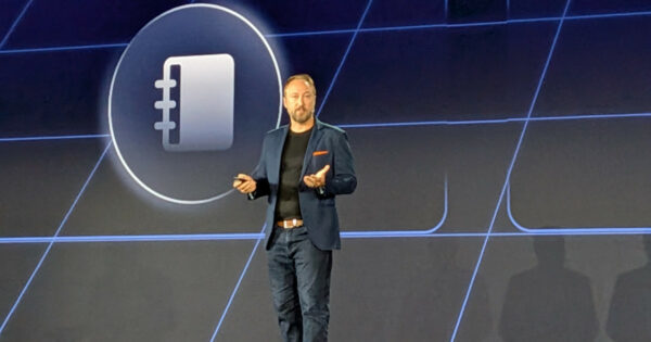 Glenn Nethercutt, CTO e Technical Fellow da Genesys, em pé no palco durante apresentação, usando blazer azul, camiseta preta e jeans, com um controle remoto na mão; ao fundo, telão com ícone de agenda em destaque. (automação)