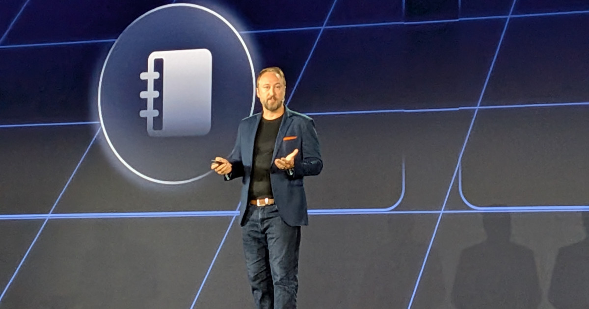 Glenn Nethercutt, CTO e Technical Fellow da Genesys, em pé no palco durante apresentação, usando blazer azul, camiseta preta e jeans, com um controle remoto na mão; ao fundo, telão com ícone de agenda em destaque. (automação)