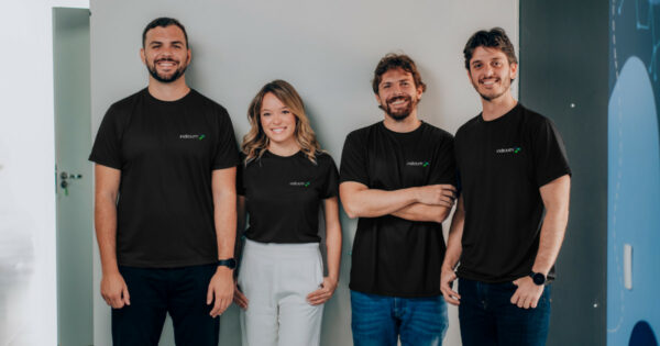 Quatro cofundadores da Indicium posam sorridentes lado a lado em frente a uma parede clara. Todos vestem camisetas pretas com o logotipo da empresa; a única mulher do grupo usa calça branca, enquanto os três homens usam calças escuras (databricks)