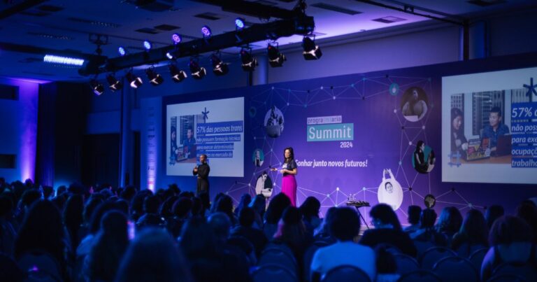 Evento PrograMaria Summit com público sentado assistindo a dois palestrantes em um palco iluminado com luzes azuis. O fundo do palco exibe os dizeres "Summit 2024" e "Sonhar junto novos futuros!". Duas telas grandes mostram o mesmo slide com a informação: "57% das pessoas trans não possuem qualificação para exercer ocupações formais de trabalho."