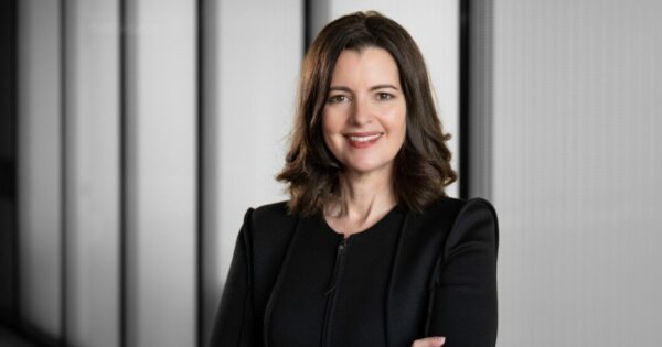 Pessoa de cabelos na altura dos ombros, vestindo roupa preta, em pé com os braços cruzados. O fundo é composto por painéis verticais em tons de cinza. Sandra Joyce, vice presidente global de Ameaças Cibernéticas da Google Cloud