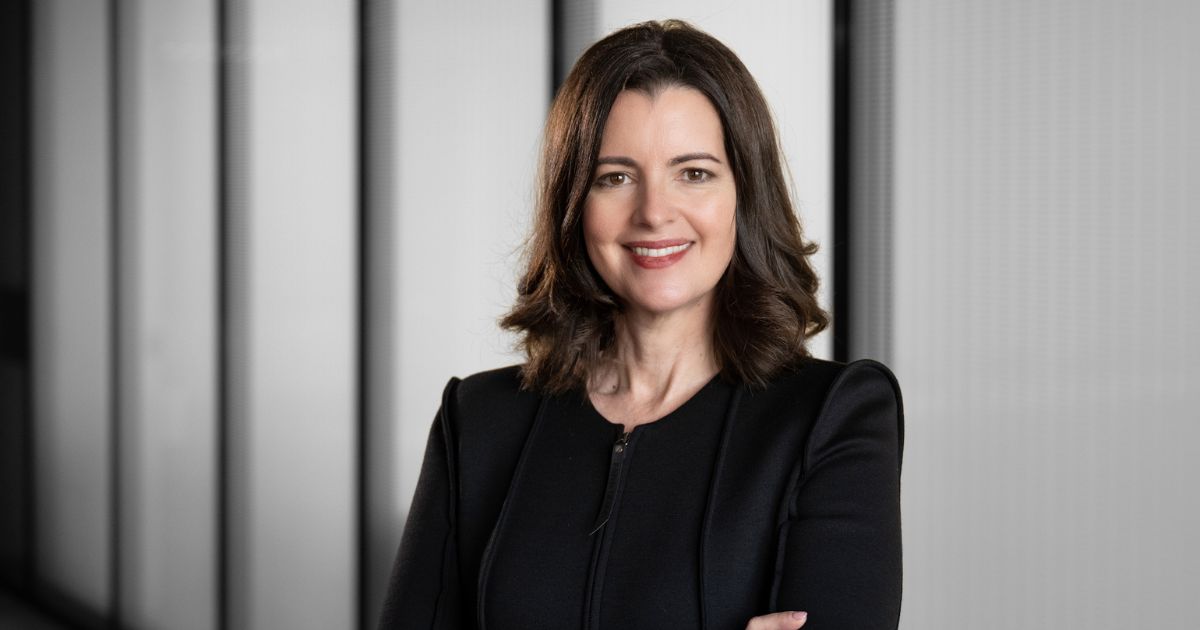 Pessoa de cabelos na altura dos ombros, vestindo roupa preta, em pé com os braços cruzados. O fundo é composto por painéis verticais em tons de cinza. Sandra Joyce, vice presidente global de Ameaças Cibernéticas da Google Cloud