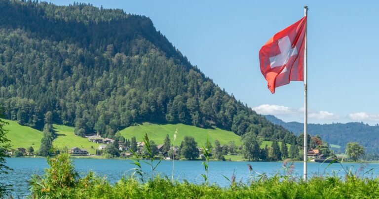 Bandeira da Suíça tremulando em um mastro à beira de um lago, com montanhas verdes cobertas de árvores e pequenas casas ao fundo em um dia ensolarado