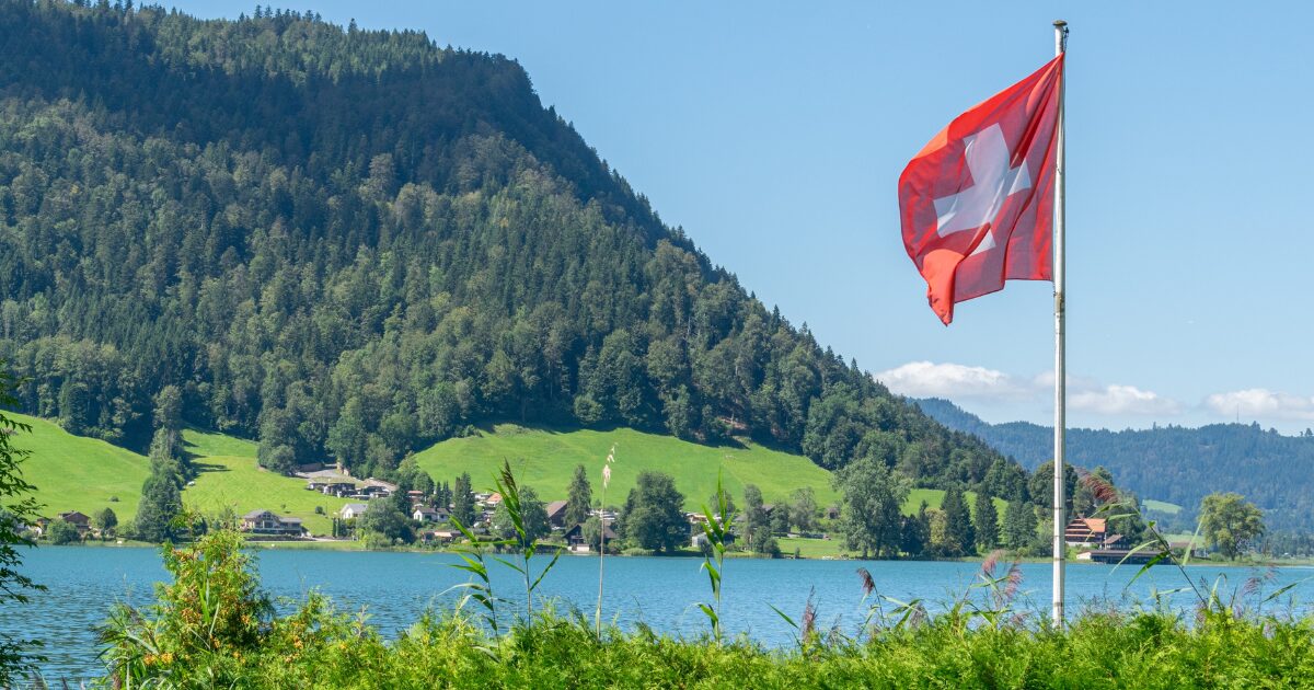 Bandeira da Suíça tremulando em um mastro à beira de um lago, com montanhas verdes cobertas de árvores e pequenas casas ao fundo em um dia ensolarado