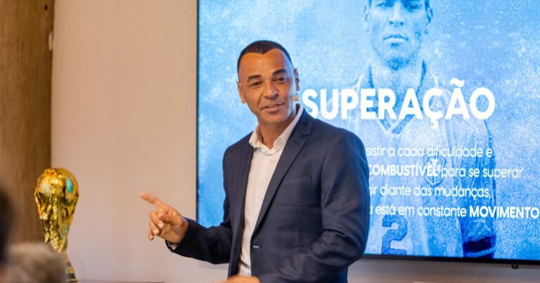 Ex-jogador Cafu, vestindo terno escuro e camisa branca, aponta para telão azul com o título "Superação" durante palestra empresarial. Na mesa ao lado, replica da taça da Copa do Mundo dourada. O ambiente é corporativo, com parede bege ao fundo (liderança)