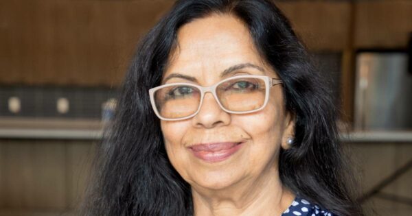 Mulher de meia-idade, de cabelos longos e pretos, usando óculos de armação clara e blusa azul com estampas brancas, sorrindo em ambiente interno. Trata-se da Dra. Sambhavi Chandrashekar, líder global em acessibilidade na D2L e vice-presidente do Comitê Técnico de Sistemas de IA Acessíveis e Igualitários da agência Accessibility Standards.
