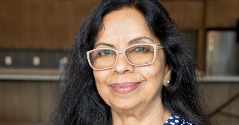 Mulher de meia-idade, de cabelos longos e pretos, usando óculos de armação clara e blusa azul com estampas brancas, sorrindo em ambiente interno. Trata-se da Dra. Sambhavi Chandrashekar, líder global em acessibilidade na D2L e vice-presidente do Comitê Técnico de Sistemas de IA Acessíveis e Igualitários da agência Accessibility Standards.