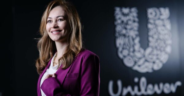 Mulher sorridente com cabelos castanhos claros, vestindo blazer roxo e blusa branca, em frente a uma parede preta com o logotipo da Unilever em destaque. A legenda informa que é Claudia Meira, CIO Latam da Unilever.