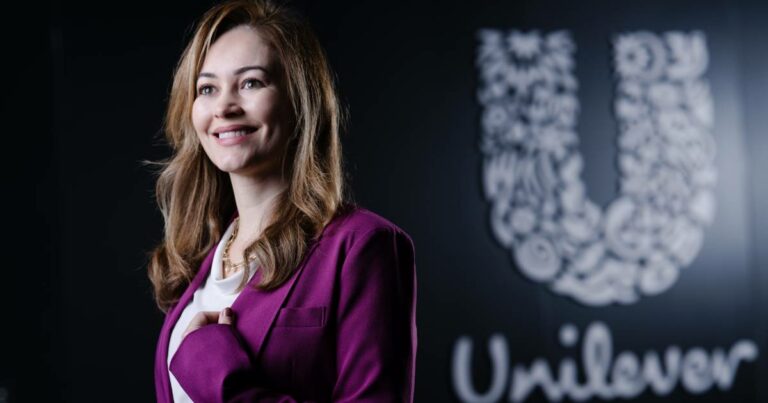 Mulher sorridente com cabelos castanhos claros, vestindo blazer roxo e blusa branca, em frente a uma parede preta com o logotipo da Unilever em destaque. A legenda informa que é Claudia Meira, CIO Latam da Unilever.