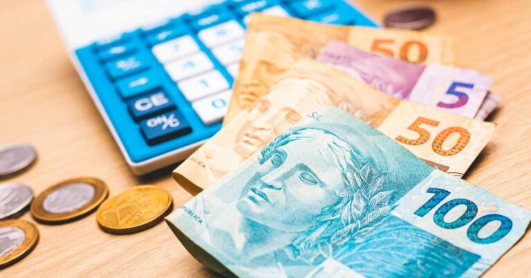 Notas de real (R$100, R$50 e R$5) e moedas espalhadas sobre uma mesa de madeira, ao lado de uma calculadora azul e branca. Representação de serviços financeiros, cálculos econômicos e gestão de recursos, fintech, crédito, brasil