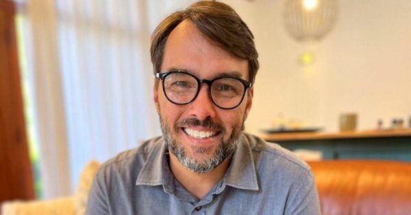 A imagem mostra uma pessoa com cabelo curto e óculos, vestindo uma camisa com gola. O fundo revela um ambiente interno com um sofá, objetos desfocados sobre uma prateleira e iluminação suave. A composição oferece uma visão clara do rosto e parte superior do corpo da pessoa em um contexto casual.
