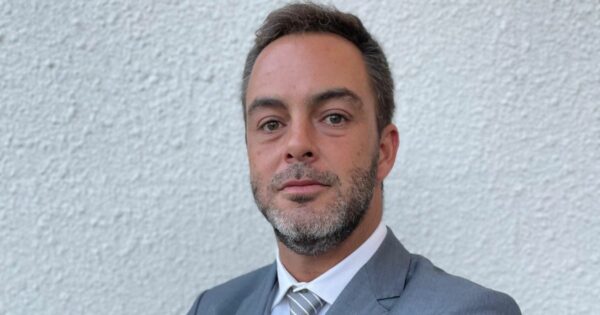 A imagem mostra uma pessoa com cabelo curto e barba, vestindo terno e gravata. Ela está posicionada diante de uma parede branca com textura. A composição tem aparência formal, adequada para contextos profissionais ou institucionais.