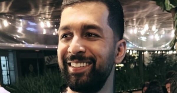 A imagem mostra uma pessoa com barba sorrindo. O fundo indica um ambiente externo, com luzes decorativas suspensas e vegetação, sugerindo um clima festivo ou descontraído.