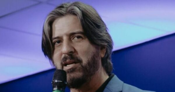 A imagem mostra uma pessoa com cabelo de comprimento médio e barba, segurando um microfone enquanto fala. O fundo apresenta um gradiente em tons de azul e roxo, sugerindo um ambiente interno com iluminação artificial. A composição indica que a pessoa está participando de um evento ou fazendo uma apresentação.