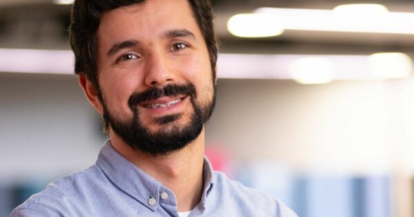 A imagem mostra uma pessoa com cabelo curto e escuro, barba e vestindo uma camisa azul clara. O fundo está desfocado, com luzes brilhantes e formas indistintas, sugerindo um ambiente interno como um escritório ou espaço de trabalho. A composição transmite um contexto profissional.