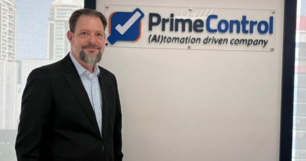 A imagem mostra uma pessoa em traje social ao lado de uma placa com os dizeres "Prime Control (AI)tomation driven company". O fundo revela um ambiente de escritório com grandes janelas que mostram uma paisagem urbana. A composição sugere um contexto corporativo ligado à empresa Prime Control, voltada para automação com uso de inteligência artificial.
