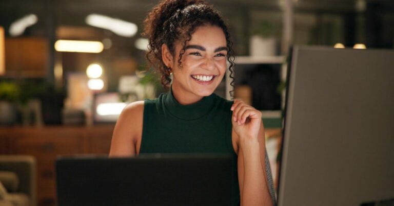 A cena mostra uma pessoa sentada em frente a uma mesa de trabalho com dois dispositivos visíveis: um laptop aberto à frente e um monitor grande à direita. A pessoa está usando uma blusa sem mangas de cor verde escura e tem cabelos cacheados presos em um coque alto, com alguns fios soltos. O ambiente parece ser um escritório moderno, com iluminação suave e elementos desfocados ao fundo, como móveis e plantas. A mão direita da pessoa está levemente levantada, próxima ao rosto, sugerindo que está pensando ou interagindo com algo na tela. (emprego, profissional)