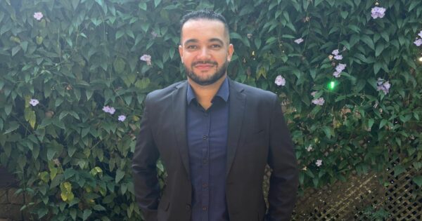 A imagem mostra uma pessoa em pé diante de um fundo verde vibrante, composto por folhas e pequenas flores roxas. Ela veste um blazer escuro sobre uma camisa também escura e está voltada diretamente para a câmera. O cenário parece ser ao ar livre, como um jardim ou parque, oferecendo um contraste natural à apresentação formal da pessoa. (startup, IA)