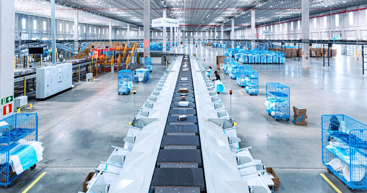 Vista ampla docentro de distribuição logístico, da Loggi com esteiras automatizadas ao centro transportando encomendas. Ao redor, há várias gaiolas metálicas azuis organizadas e funcionários trabalhando em meio ao fluxo de pacotes. O galpão é amplo, bem iluminado e estruturado com pilares altos, corredores largos e equipamentos industriais.