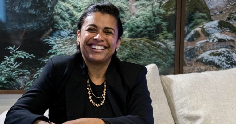 Mulher sorridente sentada em um sofá claro, usando blazer preto e colar dourado, com um painel de paisagem natural ao fundo; junto à imagem aparece o texto “Noéle Gomes, pesquisadora”. (filosofia)