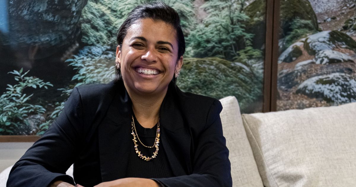 Mulher sorridente sentada em um sofá claro, usando blazer preto e colar dourado, com um painel de paisagem natural ao fundo; junto à imagem aparece o texto “Noéle Gomes, pesquisadora”. (filosofia)